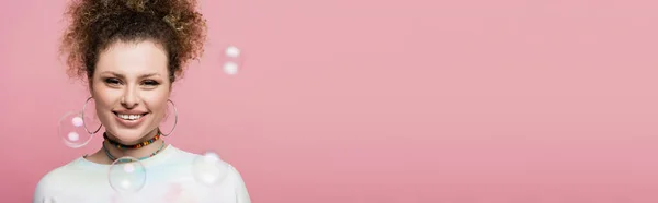 Femme positive regardant la caméra près de bulles de savon floues sur fond rose, bannière — Photo de stock
