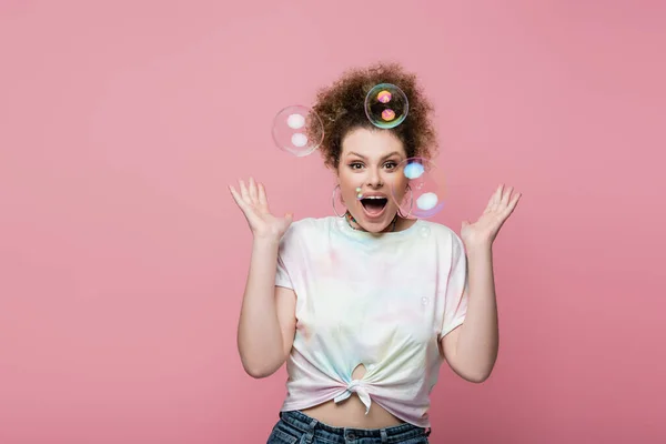 Shocked young woman standing near soap bubbles on pink background — Stock Photo