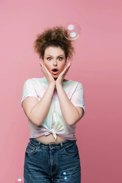 Schockierte Frau in Jeans und T-Shirt blickt in die Kamera neben Seifenblasen auf rosa Hintergrund — Stockfoto