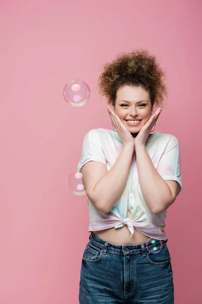 Donna positiva in t-shirt posa vicino bolle di sapone su sfondo rosa — Foto stock