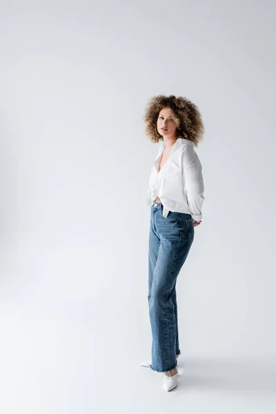 Femme bouclée en chemisier et jeans regardant la caméra sur fond blanc — Photo de stock