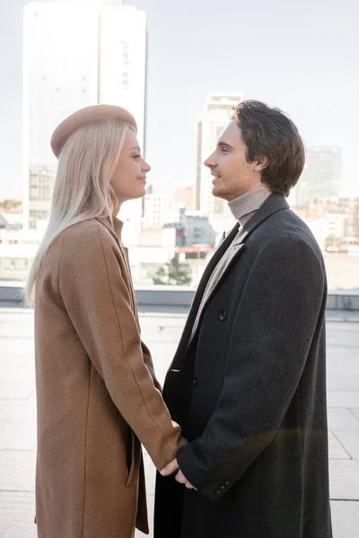 Side view of young couple in coats holding hands and looking at each other outdoors — Stock Photo