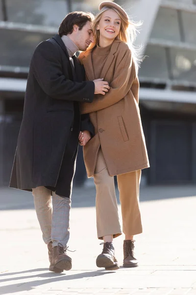 Full length of happy couple in coats and leather boots holding hands and hugging while walking on street — Stock Photo