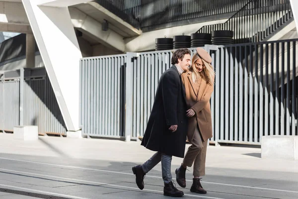 Comprimento total de casal elegante e feliz em casacos andando na rua urbana — Fotografia de Stock