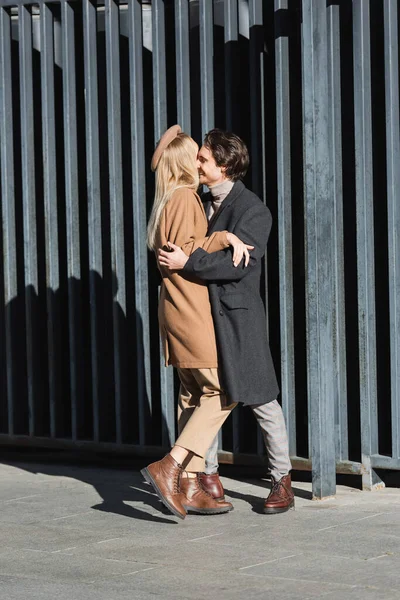 Full length of couple in leather boots and coats embracing near fence on street, side view — Stock Photo