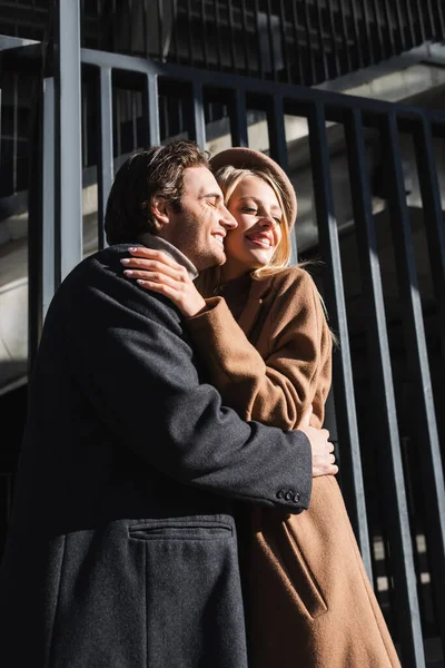 Complacida pareja en abrigos abrazando con los ojos cerrados cerca de valla - foto de stock