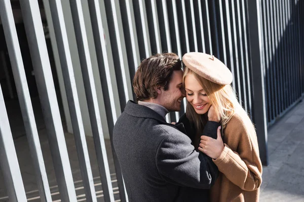 Young man embracing cheerful woman in beret near fence outdoors — Stock Photo