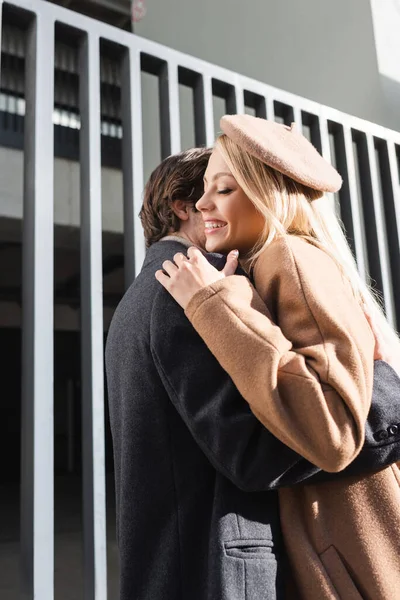 Cheerful woman in beret embracing with boyfriend near fence on street — Stock Photo