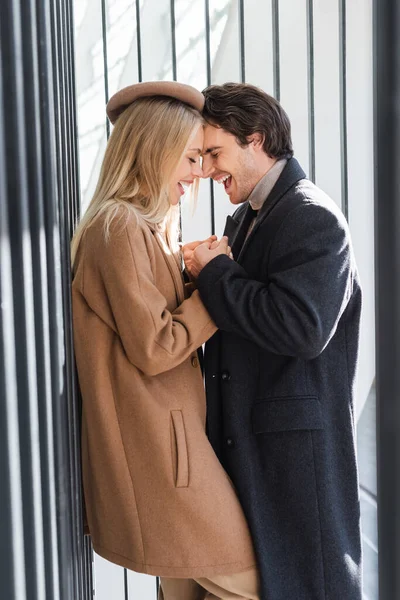 Side view of trendy couple in autumn clothes holding hands and laughing near fence — Stock Photo