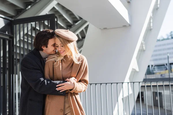 Щасливий чоловік обіймає жінку в осінньому пальто і береті біля паркану і будівлі — стокове фото