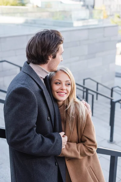 Cheerful blonde woman looking away near young man in coat outdoors — Stock Photo