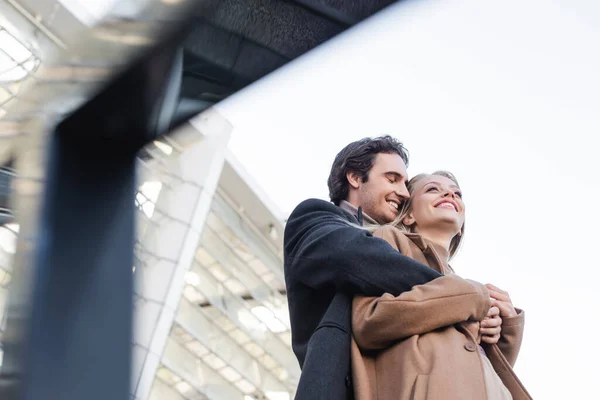 Baixo ângulo vista de homem no casaco abraçando namorada feliz em primeiro plano borrado — Fotografia de Stock