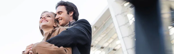 Low angle view of man with closed eyes embracing cheerful blonde woman outdoors, banner — Stock Photo