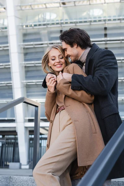 Alegre hombre en abrigo abrazando joven y elegante novia en la calle urbana - foto de stock