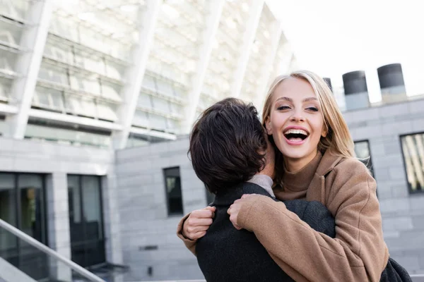 Excited blonde woman embracing boyfriend and laughing on city street — Stock Photo