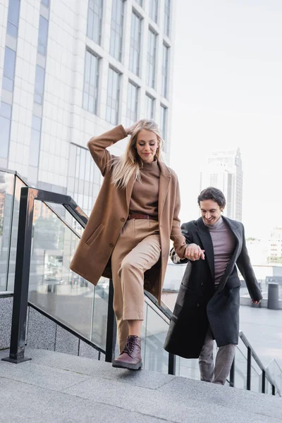 Mulher na moda no casaco de mãos dadas com o jovem enquanto caminha na cidade — Fotografia de Stock
