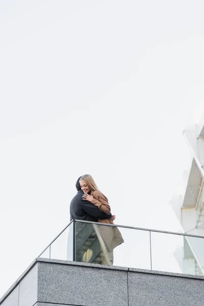 Baixo ângulo vista de mulher feliz em roupas de outono abraçando com o homem perto de cerca de vidro — Fotografia de Stock