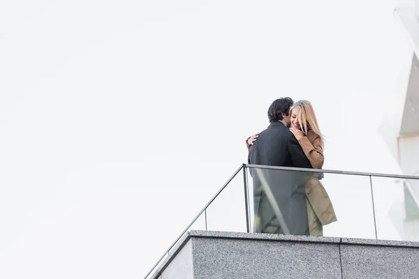 Vista de ángulo bajo de la pareja en el traje de otoño abrazándose cerca de la cerca de vidrio - foto de stock