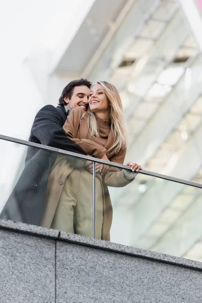 Baixo ângulo de visão do homem sorridente abraçando mulher feliz e elegante perto de cerca de vidro — Fotografia de Stock