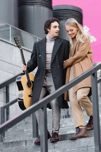 Full length of stylish man with acoustic guitar walking on stairs near cheerful blonde woman — Stock Photo