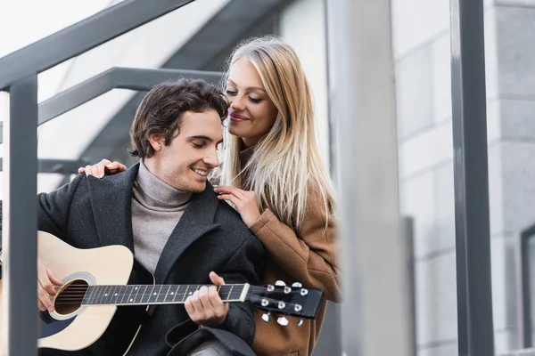 Mujer rubia sonriendo y abrazando hombros de hombre joven tocando la guitarra acústica al aire libre - foto de stock