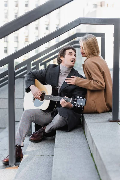 Glückliche blonde Frau in Herbstkleidung rührt Mann an, der auf Treppe Akustikgitarre spielt — Stockfoto