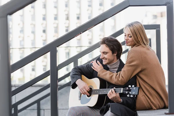 Femme heureuse étreignant jeune homme jouant de la guitare acoustique dans la rue urbaine — Photo de stock