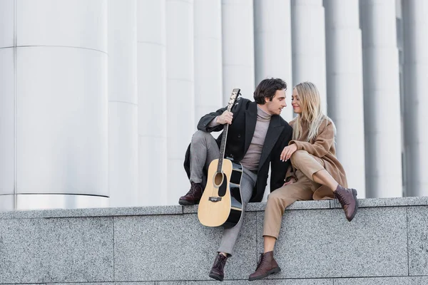 Piena lunghezza della donna in abito autunno alla moda seduto sul parapetto vicino all'uomo con la chitarra — Foto stock