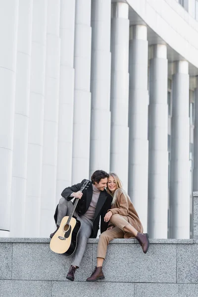 Vista completa de pareja alegre en traje de otoño elegante sentado con guitarra acústica en parapeto - foto de stock