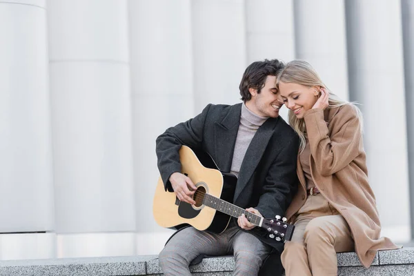 Hombre en abrigo de otoño tocando la guitarra acústica cerca de la mujer feliz mientras está sentado en el parapeto - foto de stock