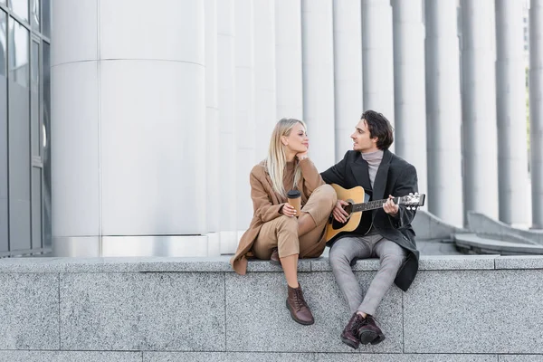Полный рост мужчины, играющего на акустической гитаре рядом с улыбающейся женщиной с кофе, чтобы пойти, сидя на парапете — стоковое фото