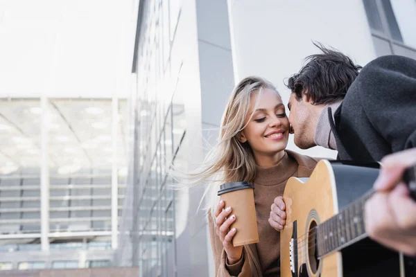 Jovem com guitarra acústica beijando mulher loira feliz com café para ir na rua da cidade — Fotografia de Stock