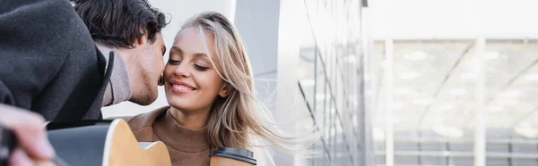 Uomo con chitarra sfocata baciare felice donna bionda con caffè per andare, banner — Foto stock