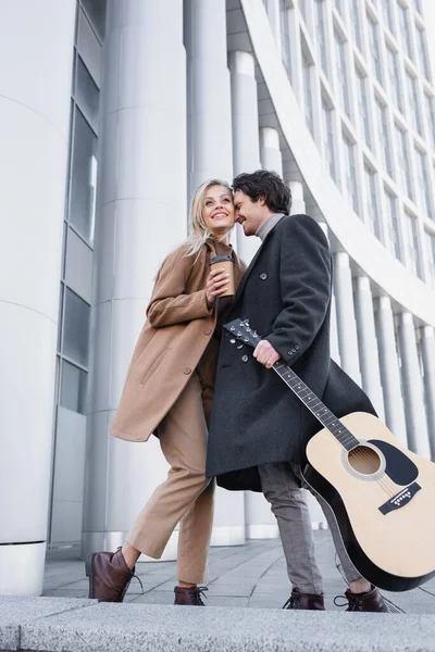 Низький кут зору пари в пальто, що стоїть з акустичною гітарою і кавою, щоб підійти до будівлі — стокове фото