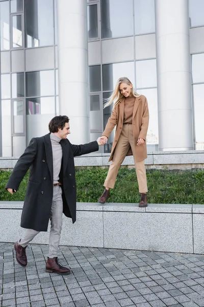 Longitud completa del hombre de abrigo cogido de la mano de la mujer de pie sobre el parapeto - foto de stock