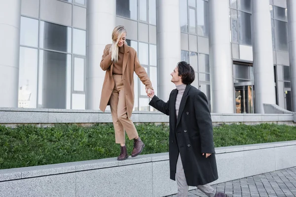 Blonde woman in autumn coat holding hands with boyfriend while walking on parapet — Stock Photo