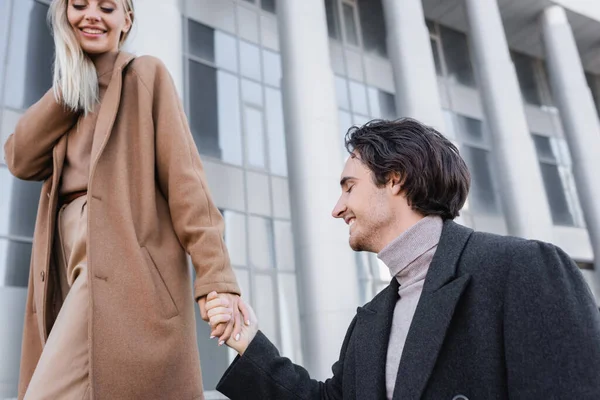 Hombre joven cogido de la mano con la mujer feliz en abrigo de moda al aire libre - foto de stock