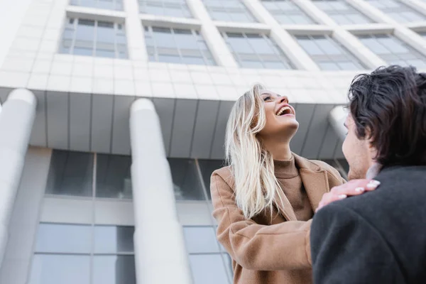 Vista de ángulo bajo de la mujer rubia riendo cerca del hombre en la ciudad - foto de stock