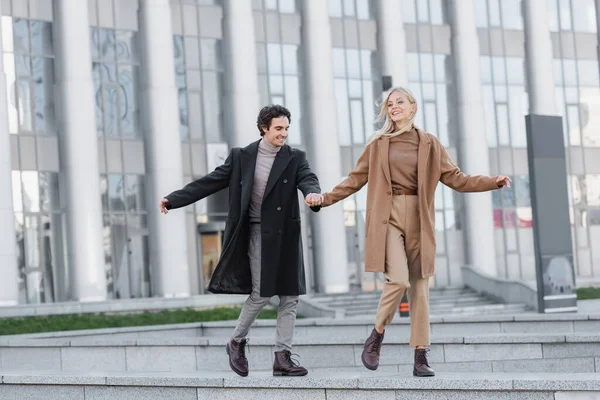 In voller Länge aufgeregtes Paar in Mänteln und Lederstiefeln, das Händchen hält und auf der Straße läuft — Stockfoto