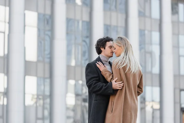 Junges Paar in Mänteln umarmt sich mit geschlossenen Augen in der Nähe des verschwommenen Gebäudes — Stockfoto