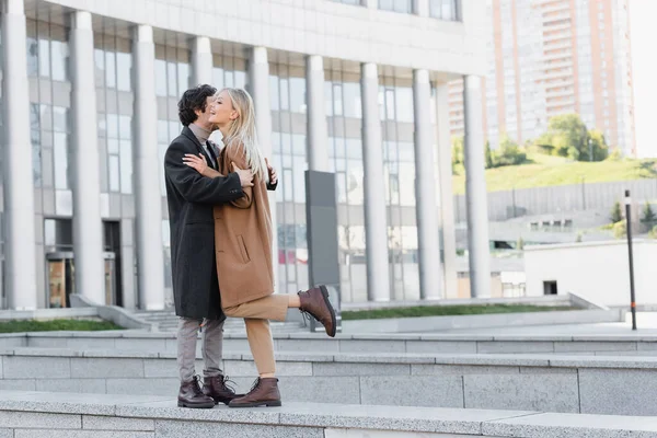 Comprimento total do casal na moda em casacos de outono e botas de couro abraçando perto de edifício borrado — Fotografia de Stock