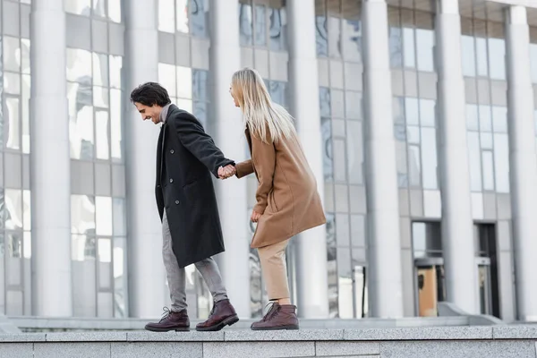 Side view of excited couple in leather boots and coats holding hands and walking on parapet — Stock Photo