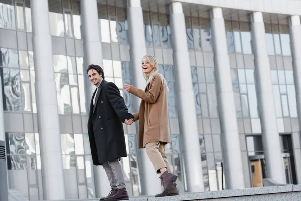 Toute la longueur du couple joyeux en bottes en cuir et manteaux d'automne tenant la main près du bâtiment flou — Photo de stock