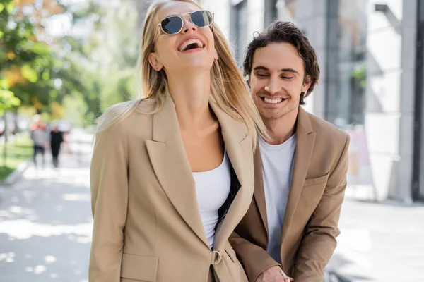 Mulher loira em óculos de sol rindo perto de homem feliz na rua urbana — Fotografia de Stock