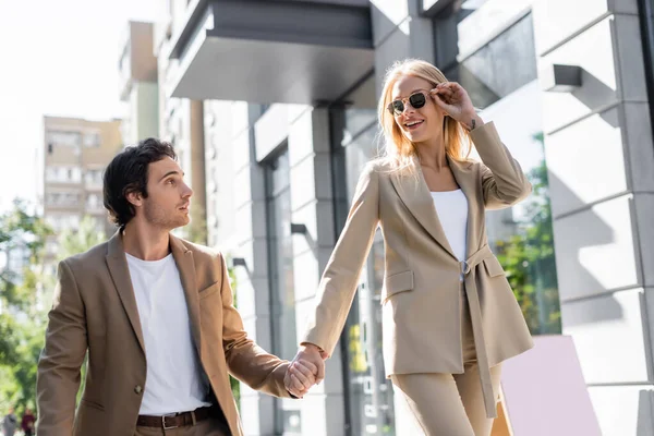 Smiling woman adjusting sunglasses while holding hands with boyfriend outdoors — Stock Photo