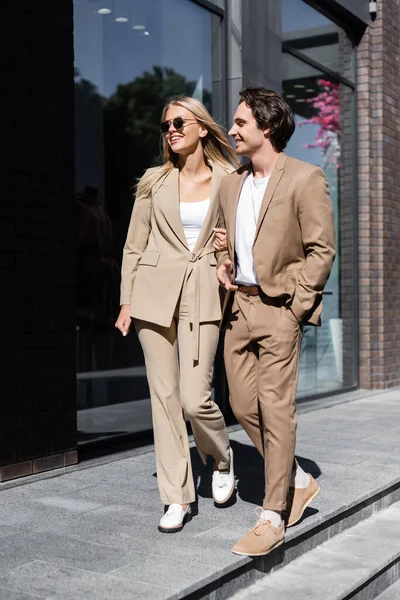 Full length of joyful young couple in stylish suits walking on urban street — Stock Photo
