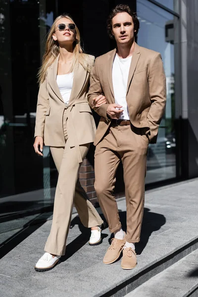 Longueur totale de couple élégant en costumes beige marchant sur la rue urbaine — Photo de stock