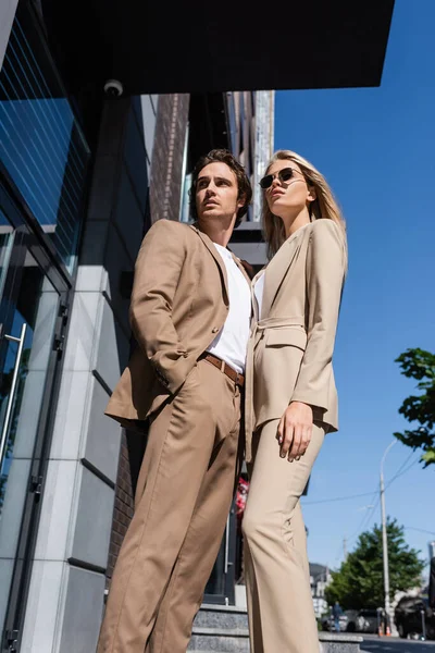 Vista de ángulo bajo del hombre con la mano en el bolsillo y la mujer rubia en gafas de sol en la calle urbana - foto de stock