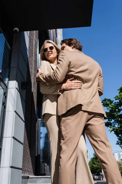 Low angle view of young couple in beige suits embracing on street in city — Stock Photo