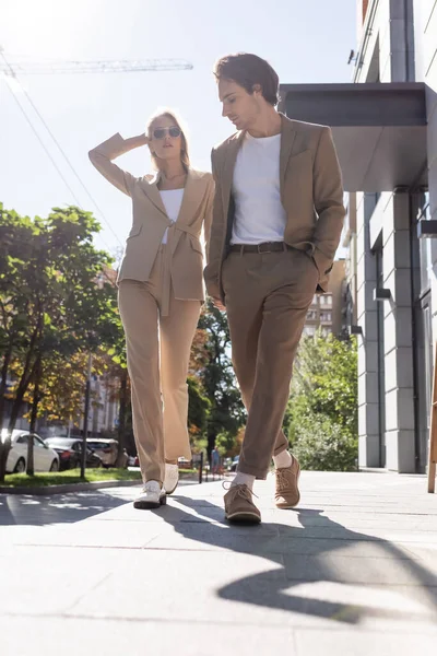 Vista completa de pareja joven en trajes beige caminando en la ciudad - foto de stock